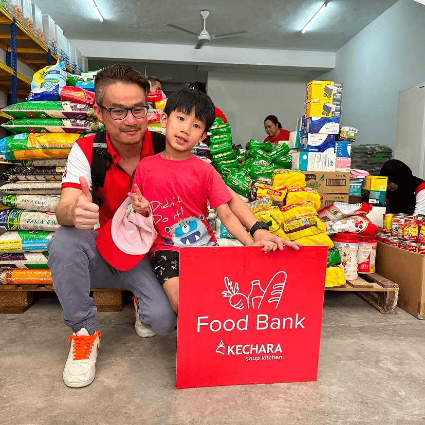 A volunteer with his kid holding a Food Bank programme flag showing thumbs up in a KSK warehouse